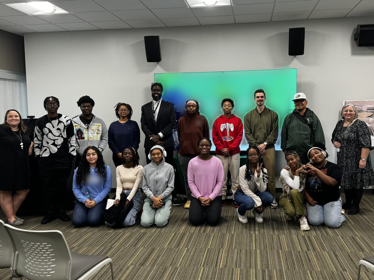 A large group of students and several adults posed together in a carpeted campus meeting room, one row standing and one row kneeling in front, with a lit display screen behind them.