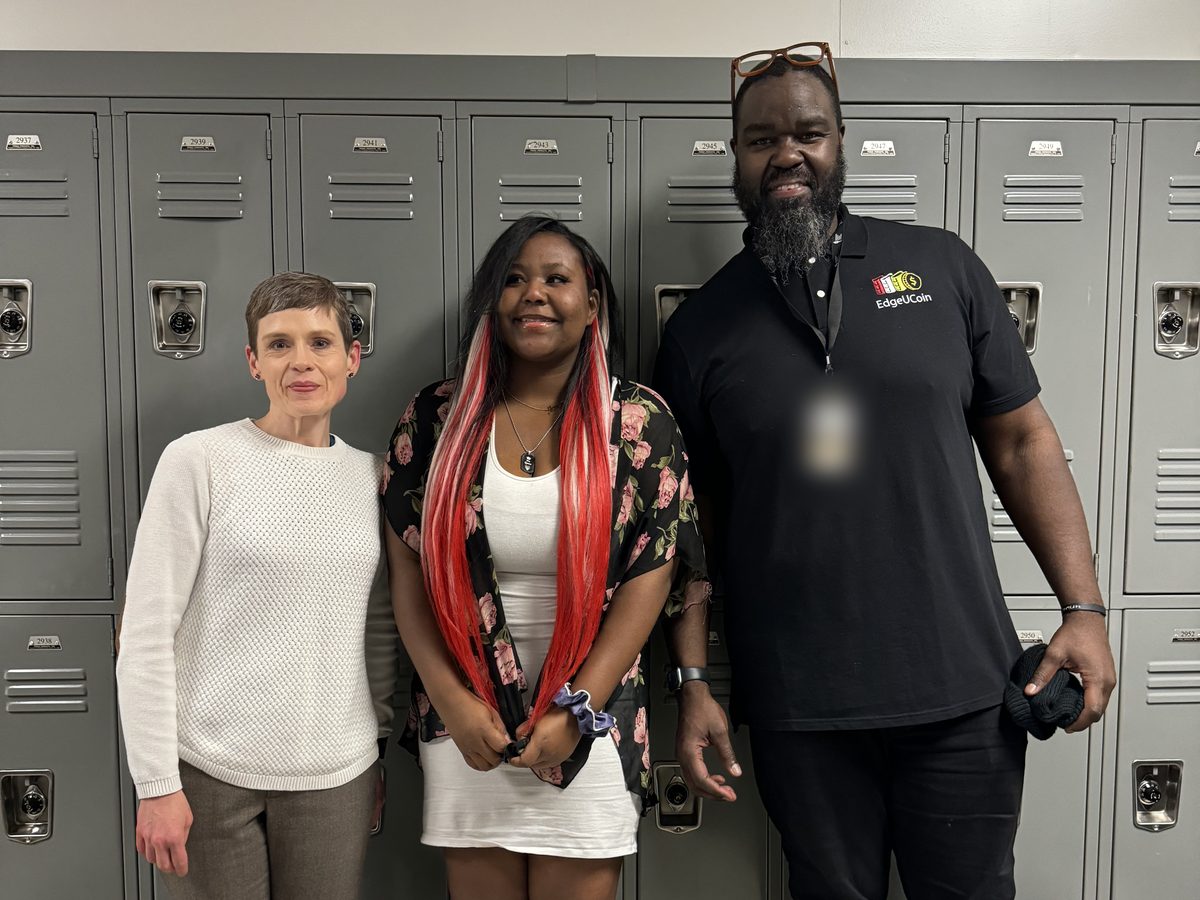Three people stand together in front of a wall of gray school lockers: a woman in a cream sweater, a teenage girl in a white dress and a floral wrap, and a bearded man in a black EdgeUCoin collared shirt.